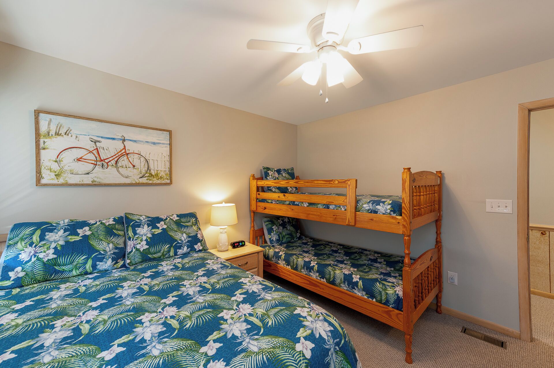 A bedroom with a bunk bed, a single bed with floral bedding, a nightstand with a lamp, a ceiling fan, and a painting of a bicycle on the wall.