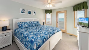 A bedroom with a bed, a nightstand, a lamp, a ceiling fan, a window with blinds, and a television. - Dolphin's Watch: A stunning oceanfront 4BR retreat in Supply, NC, perfect for 10 guests. (Supply)