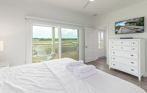 A bedroom with a large bed, a white dresser, a television, and a balcony with a view of a grassy area. - Hobbs Realty - Tidewater: Private Pool, Elevator, $250 Beach Gear Credits! (Holden Beach)