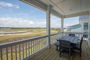 A covered porch with a table and chairs, overlooking a grassy area and a distant building. - Hobbs Realty - Tidewater: Private Pool, Elevator, $250 Beach Gear Credits! (Holden Beach)