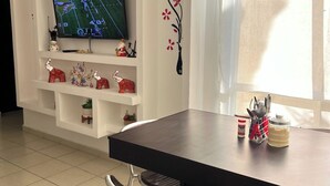 Un salon avec un téléviseur à écran plat posé sur un meuble de télévision blanc, une table à manger avec des chaises et une fenêtre avec des rideaux.