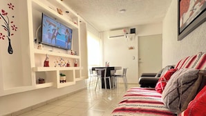 Un salon avec un téléviseur posé sur un meuble de télévision blanc, un coin repas avec une table et des chaises, et un canapé agrémenté de coussins à motifs.
