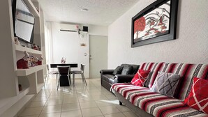 Un salon comprenant un canapé, une table à manger avec des chaises, un téléviseur fixé au mur et un ventilateur de plafond.
