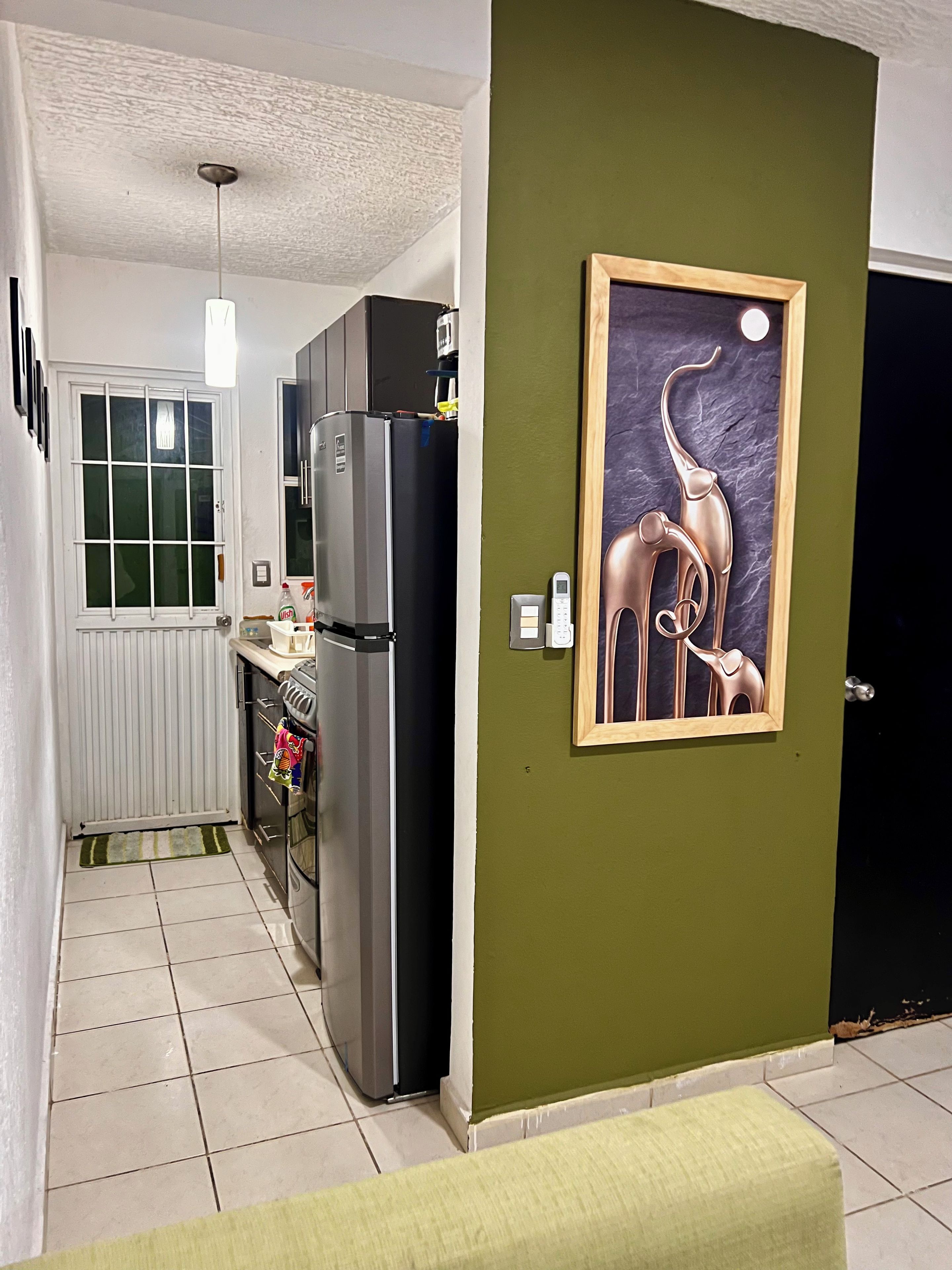 Een keuken met een koelkast, oven en een raam. Er is een deur met een foto in een lijst op de muur.