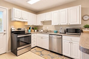 A modern kitchen with white cabinetry, stainless steel appliances, and a patterned rug. - Beach Escape • Pool View • Walk to Beach + Food (Fort Walton Beach)