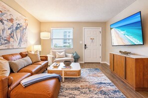 A living room with a brown leather sofa, a white armchair, a coffee table, and a flat-screen TV on the wall. - Beach Escape • Pool View • Walk to Beach + Food (Fort Walton Beach)