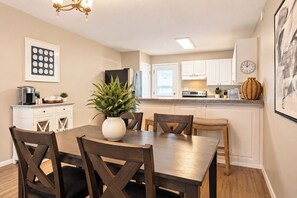 A dining area with a wooden table and chairs, a potted plant, and a kitchen in the background. - Beach Escape • Pool View • Walk to Beach + Food (Fort Walton Beach)