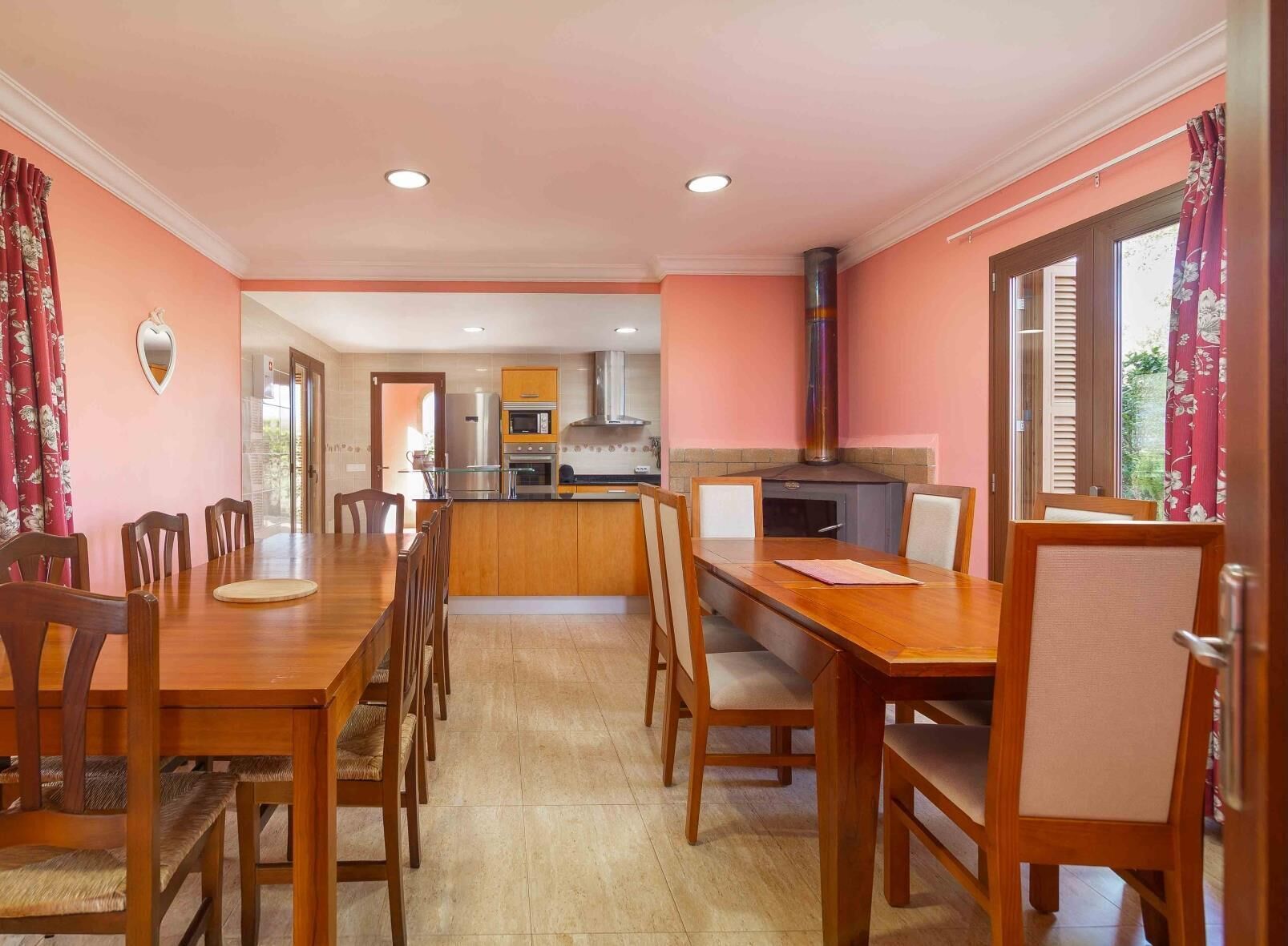 A dining area with a wooden table and chairs, a kitchen in the background, and a large window with curtains.