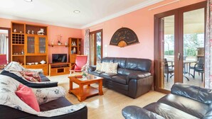 Sala de estar com sofá, mesa de centro e televisão. Há um armário de madeira com objetos decorativos e um ventilador na parede.
