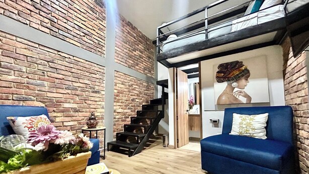 A modern living room with a brick wall, a blue sofa, a wooden coffee table with books and flowers, and a staircase leading to a loft area. - Tu Hotel Chía (Chía)