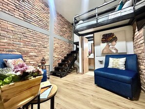 Een moderne woonkamer met een bakstenen muur, een blauwe bank, een houten salontafel met boeken en bloemen, en een trap naar een vide.