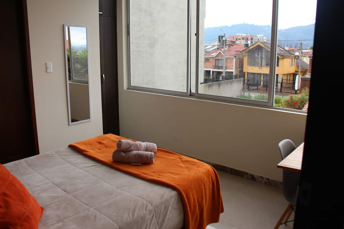 Une chambre d’hôtel avec un lit, une couverture orange, un miroir et une fenêtre donnant sur des immeubles.