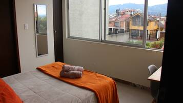 Una habitación de hotel con una cama, una manta naranja, un espejo y una ventana con vistas a edificios.