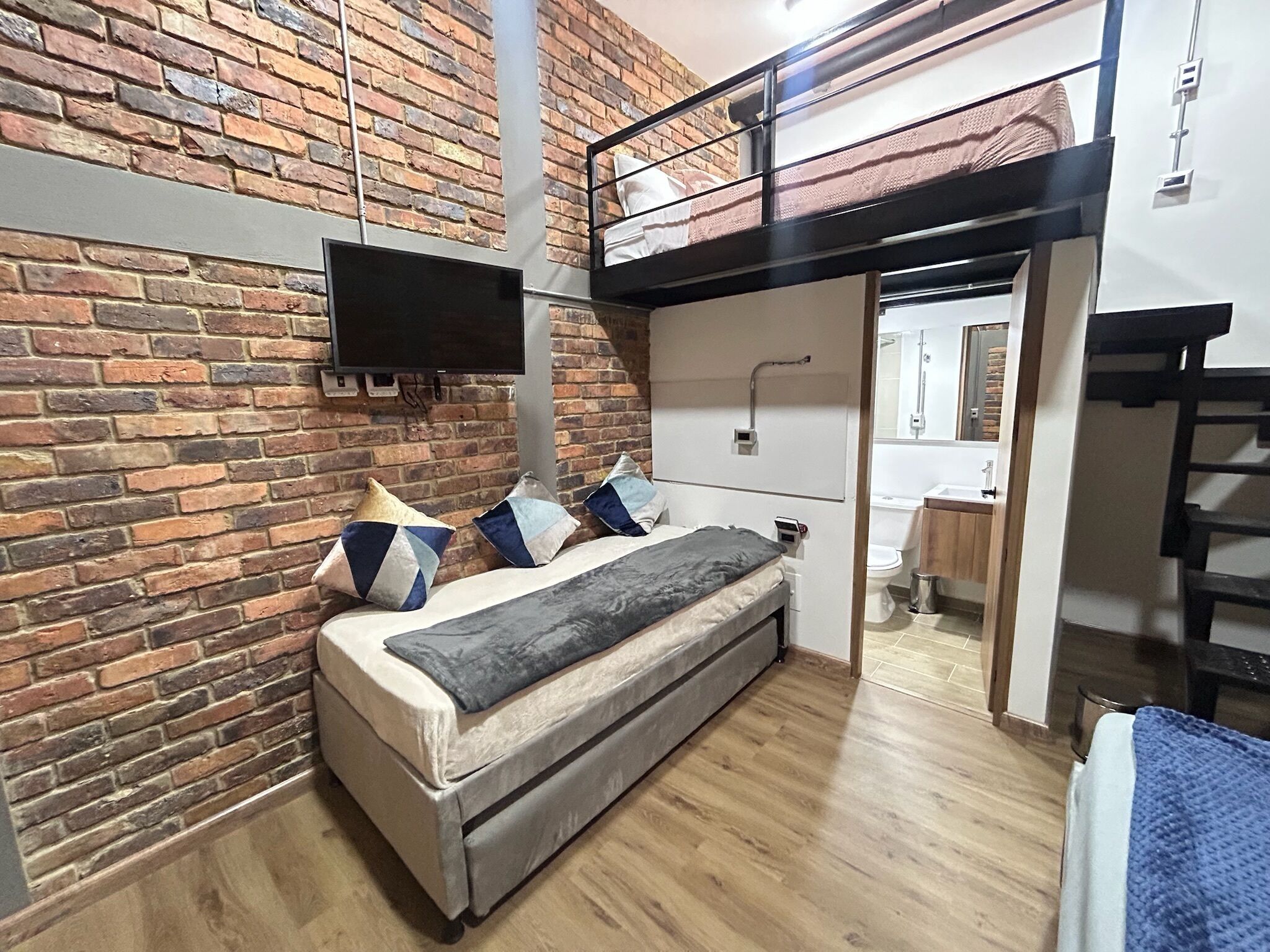 A bedroom with a loft bed, a brick wall, a flat-screen TV, and a bathroom visible through an open door.