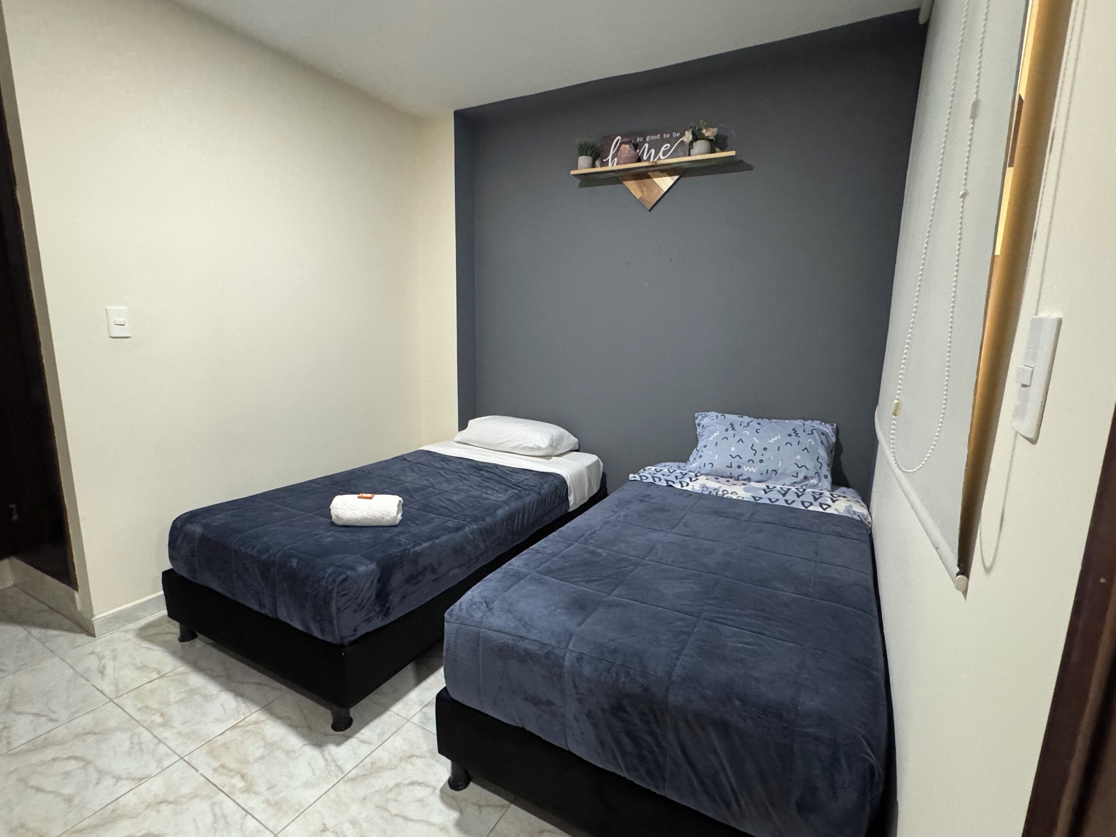 A small room with two beds, a dark wall, a shelf with decorative items, and a door.