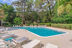 Ein Pool mit weißen Liegestühlen und einer Betonplattform, umgeben von Bäumen und viel Grün.