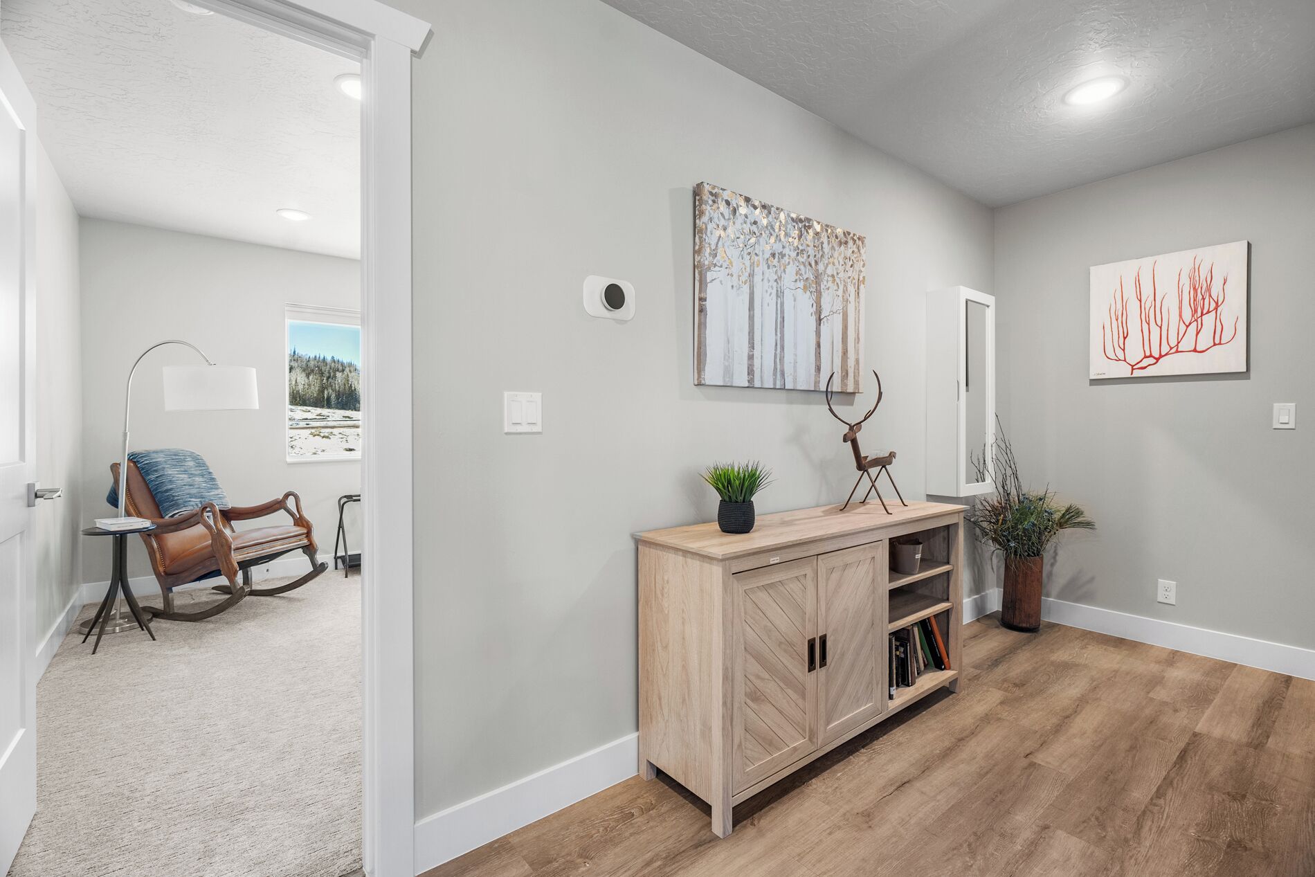 A hallway with a wooden floor, a light-colored wall, and a wooden console table with a plant and a deer sculpture. There are two framed artworks on the wall and a door leading to another room.