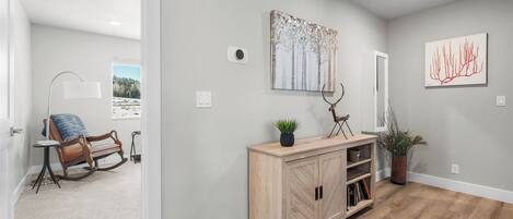 A hallway with a wooden floor, a light-colored wall, and a wooden console table with a plant and a deer sculpture. There are two framed artworks on the wall and a door leading to another room.
