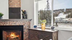 Ein Kamin mit Feuer, eine Bärenskulptur, ein Fenster mit Blick auf eine verschneite Landschaft und eine kleine Pflanze auf dem Kaminbrett.