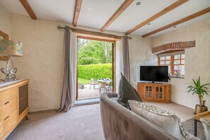 A living room with a sofa, TV, wooden cabinet, and a view of a garden through a glass door. - Cowshed East Orielton, Holiday Cottage Nr Bosherston, Sleeps 4, 2 bedrooms,  bathrooms (Pembroke)