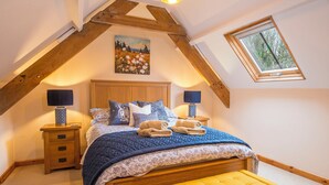 Una camera da letto con soffitto spiovente, una testiera in legno, un quadro sopra il letto, due lampade da comodino, un comodino, una panca e una finestra con vista sugli alberi.