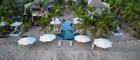 A resort with a central swimming pool, multiple thatched-roof buildings, and palm trees.