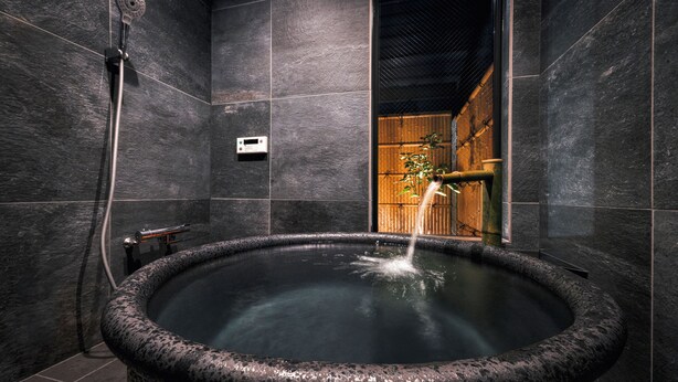 A modern bathroom with a large, circular hot tub, dark stone walls, and a wooden door leading to another room. - Rinn Kyoto Ebisunocho (Kyoto)