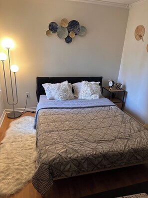 A bedroom with a bed, fluffy white rug, patterned bedspread, bedside table with a clock, and wall decor. - Queen private bedroom (Surrey)