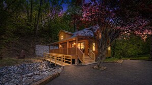 Una cabaña de troncos con una terraza de madera, un muro de piedra y un camino de entrada pavimentado.