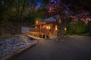 A log cabin with a wooden deck, a stone wall, and a paved driveway. - Creekfront Near Parkway | Hot Tub | Walk to Pool | 2 King Suites | Pet Friendly (Pigeon Forge)