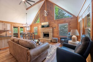 A cozy cabin interior with a fireplace, leather seating, and large windows. - Creekfront Near Parkway | Hot Tub | Walk to Pool | 2 King Suites | Pet Friendly (Pigeon Forge)
