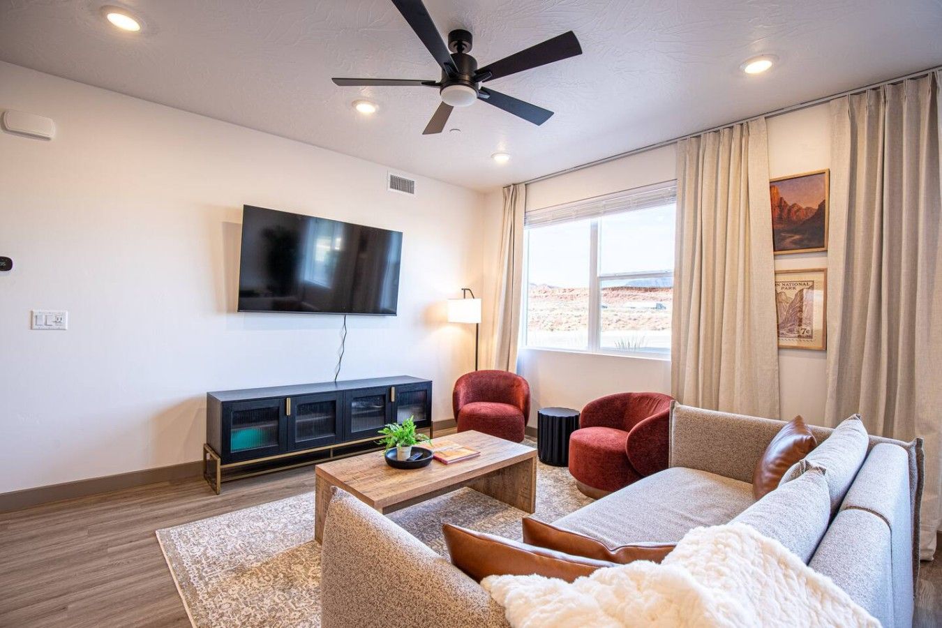 A modern living room with a flat-screen TV, a ceiling fan, a sofa, a coffee table, and a view of a desert landscape through the window.