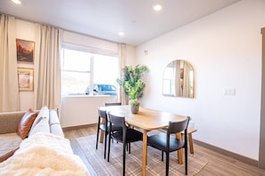 Une pièce comprenant une table à manger, des chaises, un canapé, une plante en pot, un miroir et une fenêtre avec des rideaux.
