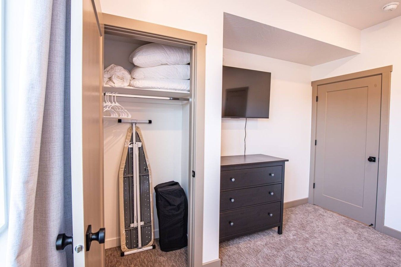 A hotel room with a closet containing folded clothes, a hanging garment, and a suitcase. There is a flat-screen TV mounted on the wall and a dark-colored dresser with drawers.
