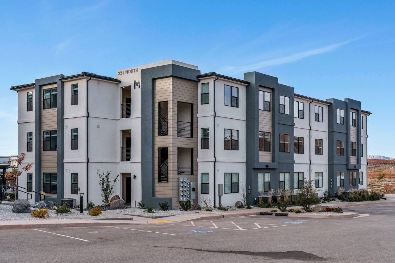 Un immeuble résidentiel à plusieurs étages, doté d’un design moderne, avec une façade combinant des couleurs blanches et grises, de grandes fenêtres et un parking situé à l’avant.