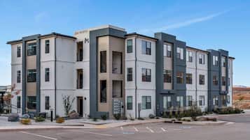 Un immeuble résidentiel à plusieurs étages, doté d’un design moderne, avec une façade combinant des couleurs blanches et grises, de grandes fenêtres et un parking situé à l’avant.
