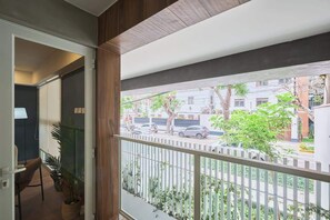Ein Balkon mit weißer Brüstung, Blick auf eine Straße mit Autos und Gebäuden, sowie eine Tür zu einem Innenraum mit einem Stuhl und einer Pflanze.