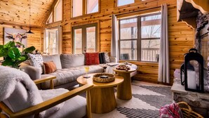 A cozy log cabin interior with a sofa, two round wooden tables, a stone fireplace, and large windows. - 5* Family Cabin~View~Hot Tub~FirePit~Pools~Bears (Gatlinburg)
