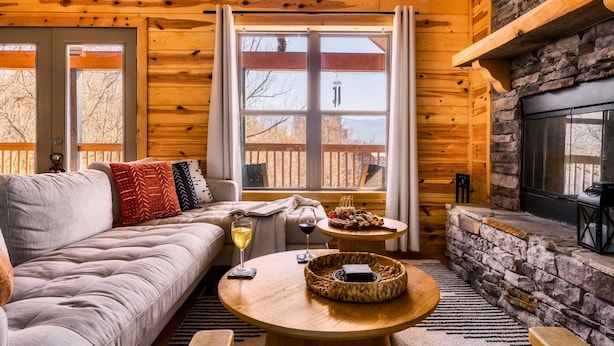 A cozy cabin interior with a wooden interior, a stone fireplace, a sofa, and a dining table with food and drinks. - 5* Family Cabin~View~Hot Tub~FirePit~Pools~Bears (Gatlinburg)