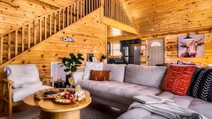 A cozy cabin interior with a wooden staircase, a living room with a sofa and coffee table, and a kitchen area. - 5* Family Cabin~View~Hot Tub~FirePit~Pools~Bears (Gatlinburg)