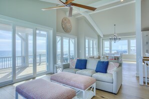 A spacious living room with a large window overlooking the ocean, a ceiling fan, and a chandelier. - Luxury Oceanfront | Saltwater pool | Elevator | Game Room | Pet Friendly (North Topsail Beach)
