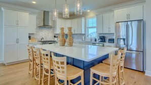A modern kitchen with white cabinets, a large island with bar stools, hanging pendant lights, and a stainless steel refrigerator. - Atlantis | Ocean Views, Private Saltwater Pool, Elevator, Pets, Beach access (Topsail Beach)