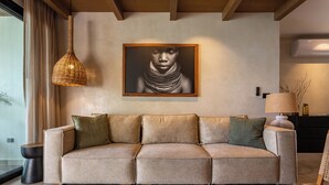 Un canapé beige agrémenté de plusieurs coussins, un luminaire suspendu en fibres tressées et une œuvre d’art encadrée accrochée au mur.