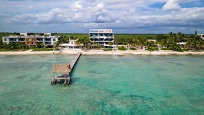 Un hôtel en bord de mer, doté d’une jetée qui s’avance dans des eaux turquoise, entouré de palmiers et surplombé d’un ciel dégagé.