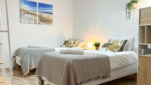 Ein Schlafzimmer mit zwei Betten, einem Nachttisch, einer Wandablage mit einer Pflanze und zwei Wandbildern mit Strandmotiven.