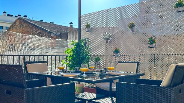 Eine Terrassengruppe im Freien mit Tisch und Stühlen aus Rattan, Platz für zwei Personen. An der Wand befinden sich Blumentöpfe und hängende Pflanzgefäße.