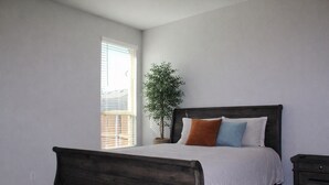 Un dormitori amb un llit de fusta, una tauleta de nit, una finestra amb persianes i una planta en test.