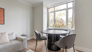Un salon moderne avec un canapé gris, une table ronde entourée de quatre chaises et une grande fenêtre donnant sur un paysage arboré.