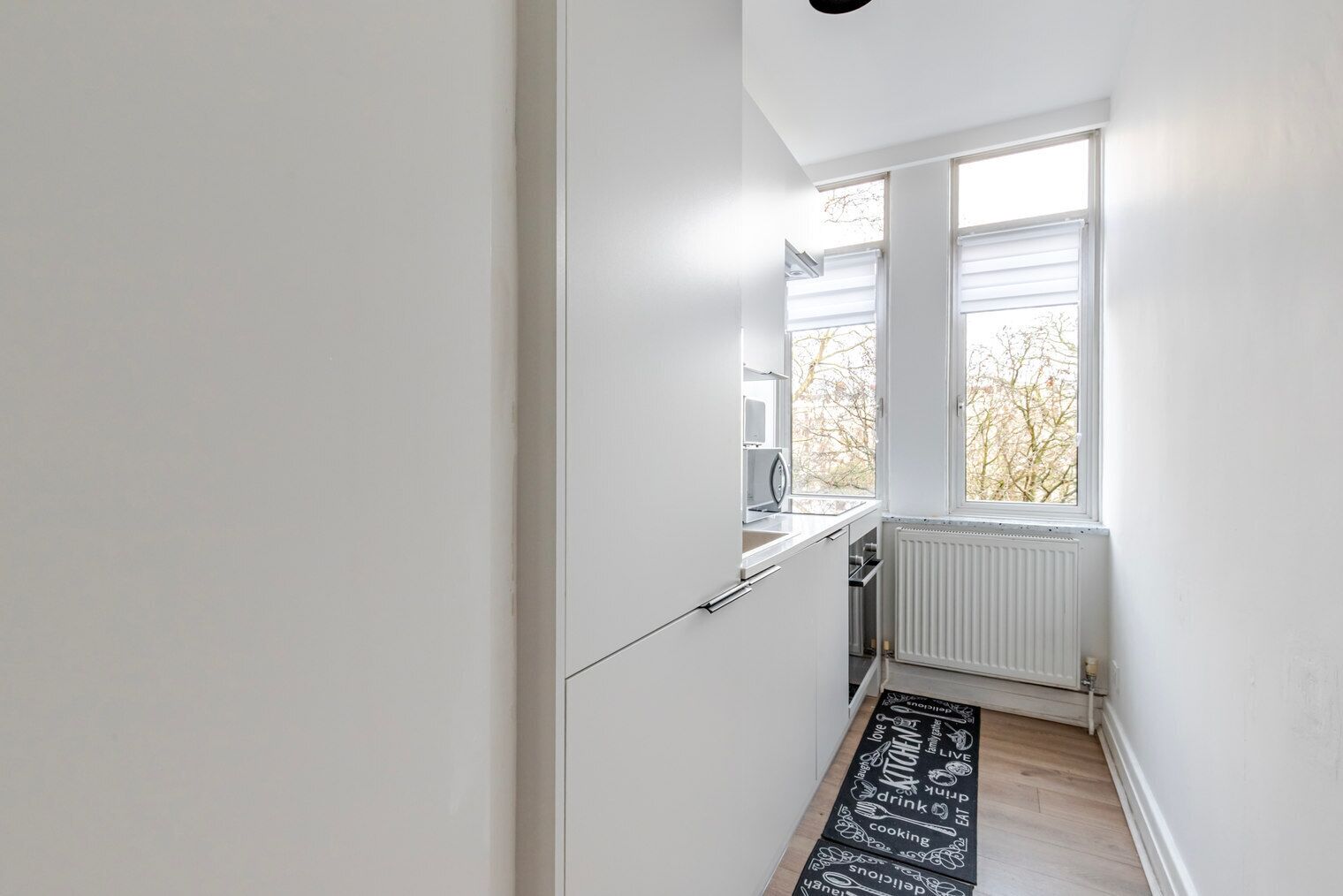 Een smalle gang met een raam en radiator, leidend naar een keuken met witte kasten en een zwarte keukenvloermat.