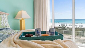 Cama com vista para a praia, uma luminária e um suporte com duas canecas e um donut.